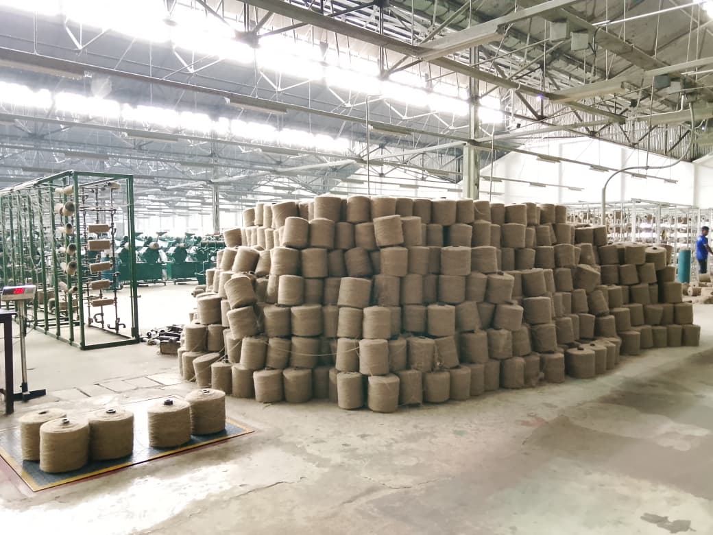 A pyramid of jute yarn cops stacked on the spinning hall floor, spinning frames and workers visible behind